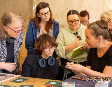 A group of people listen to an artist and look at what she is holding. Fiona Harrington, Artist in Residence at 14 Henrietta Street, explains Lace sampling to Culture Club attendees.