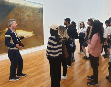 A group of people at a Culture Club listen to a guide telling them about a large painting behind them.