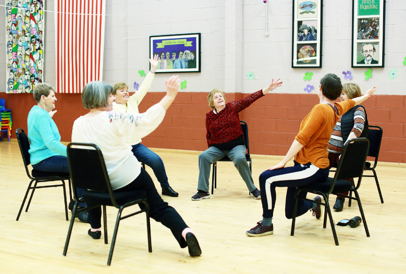 A group of woman seated on chairs extend their right arms as part of a dance class