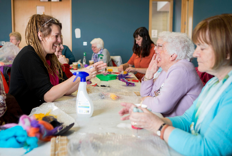 Three women converse at a table filled with craft materials as around them other people engage in arts and crafting activities