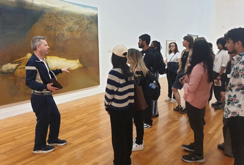 A group of people at a Culture Club listen to a guide telling them about a large painting behind them.