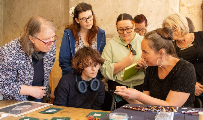 A group of people listen to an artist and look at what she is holding. Fiona Harrington, Artist in Residence at 14 Henrietta Street, explains Lace sampling to Culture Club attendees.